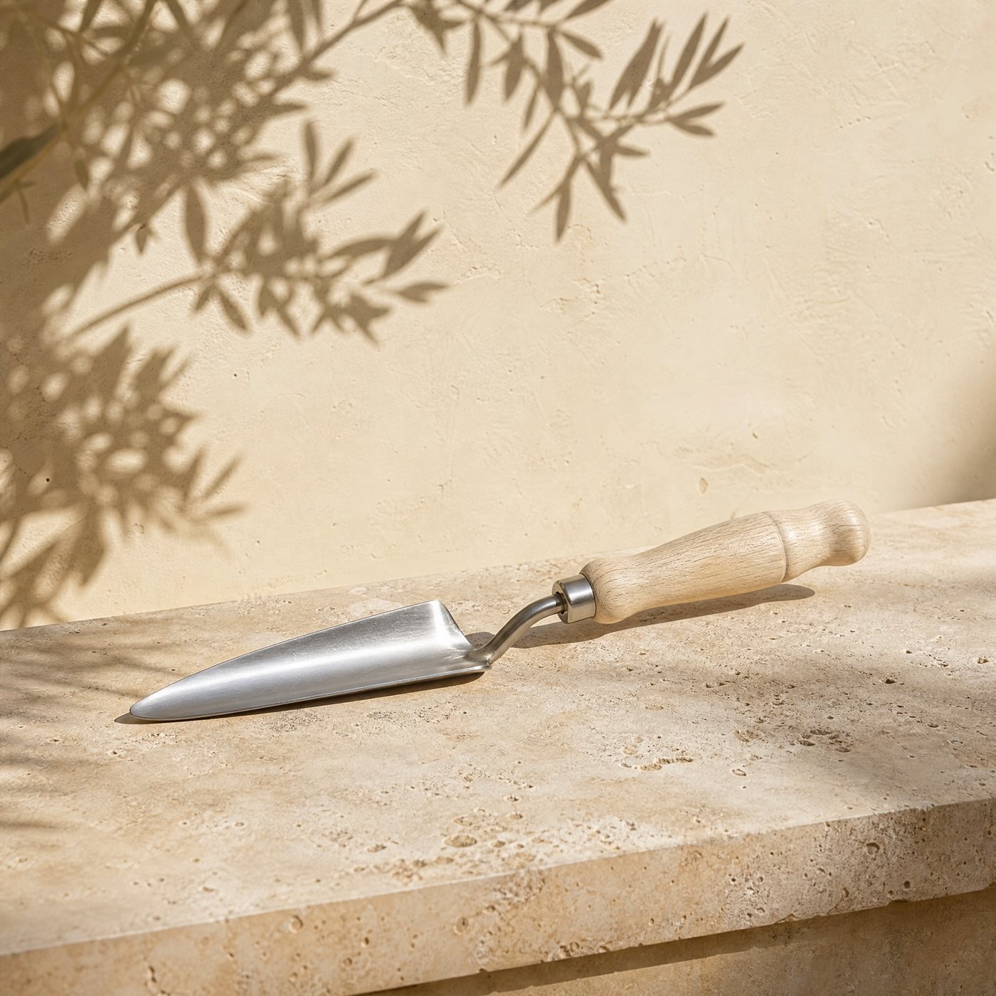 Transplanting Trowel with Depth Marks — a narrow stainless steel transplanting trowel with a turned pale ash handle, lying diagonally on a sunlit limestone slab