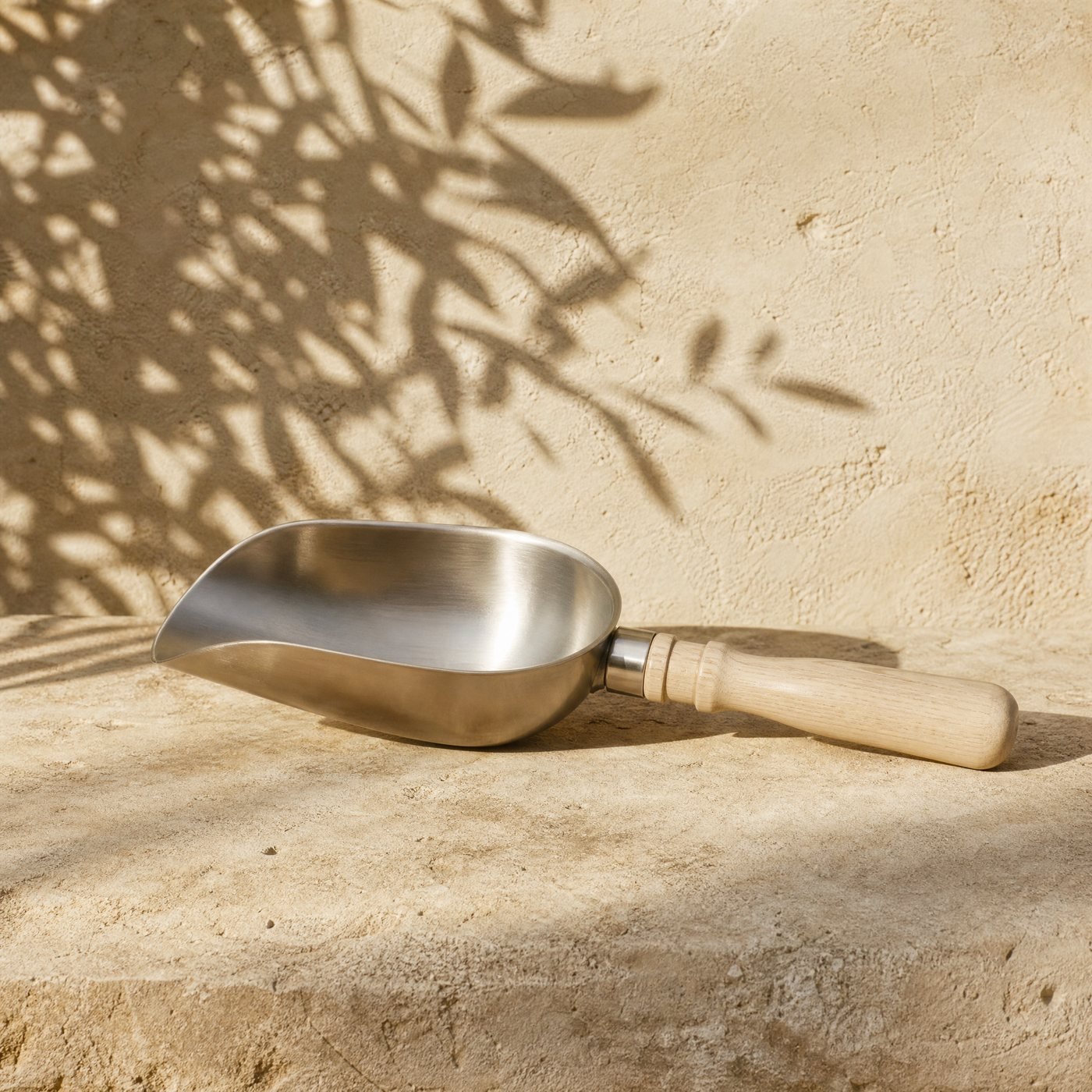 Potting Scoop, Stainless — a deep rounded stainless steel potting scoop with a short turned pale ash handle, lying on a sunlit stone slab